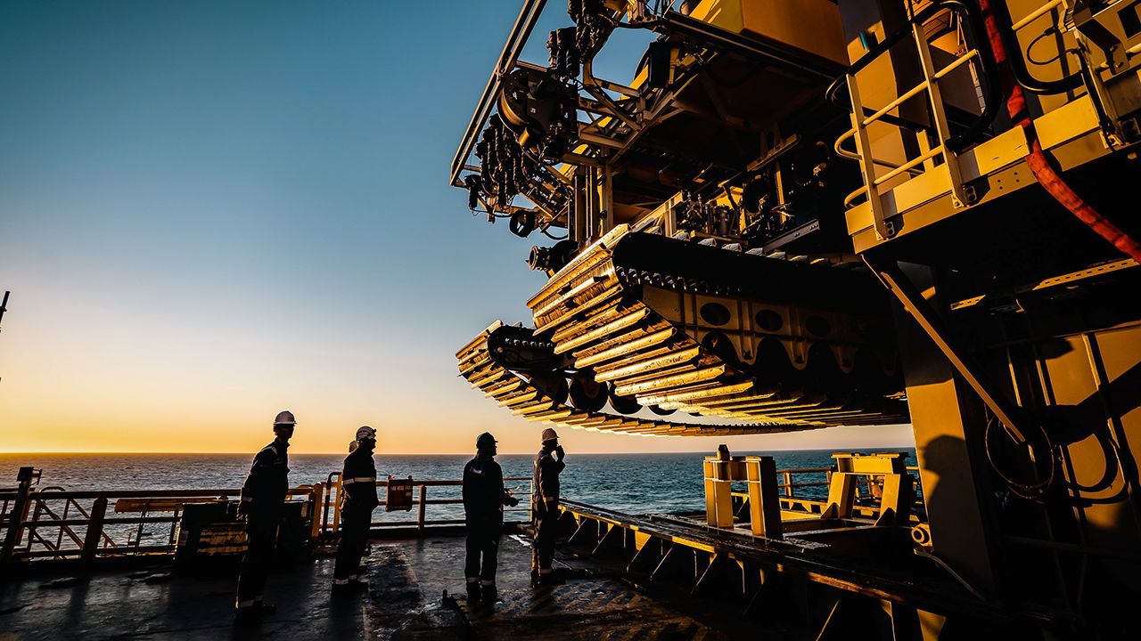 A deep sea mining nodule collector vehicle is suspended above the ocean with workers looking on.