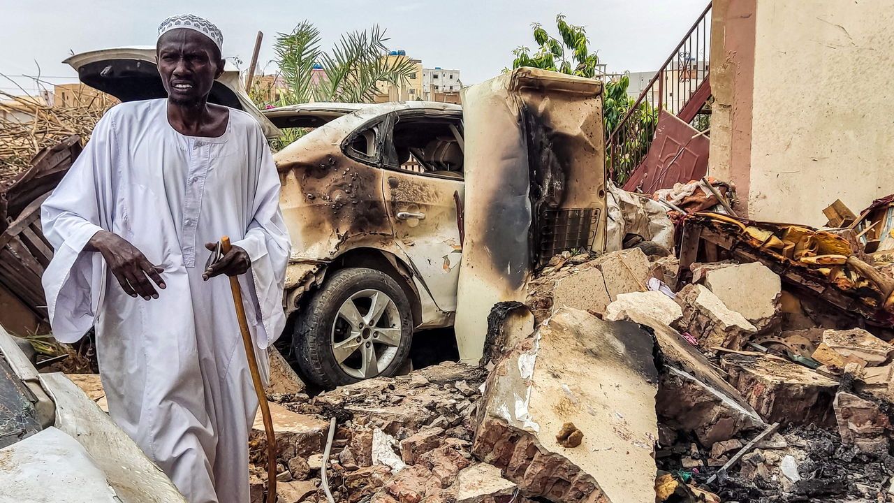 A man walks through rubble by a destroyed car and house that was hit by an artillery shell in Sudan.